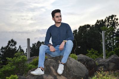 Young man looking away while sitting on rock against sky