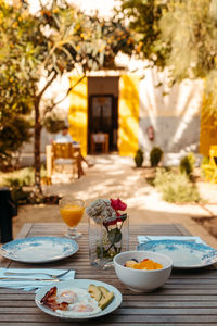 Delicious fried eggs with bacon and avocado served on wooden table with bowl of fresh fruits and glass of orange juice on sunny day in backyard of house in mallorca