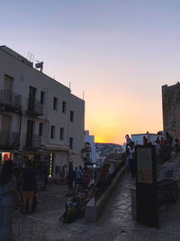 People on street amidst buildings against sky during sunset