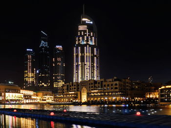 Illuminated modern buildings by river against sky at night