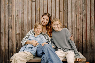 Portrait of smiling woman sitting with sons on bench in front of wooden fence