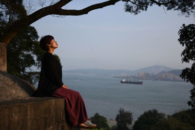 Side view of woman sitting on retaining wall against river