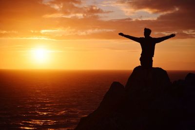 Silhouette man with arms outstretched standing on rock at beach against sky during sunset