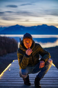 Portrait of young man crouching on pier against lake