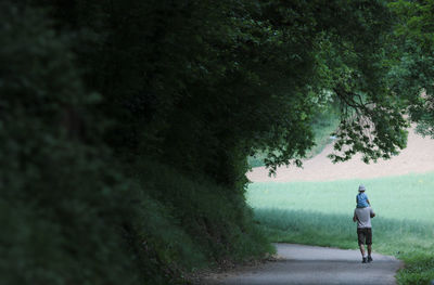 Rear view of father with daughter on shoulder walking on road amidst trees