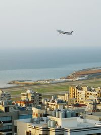 Airplane flying over sea by buildings in city against sky