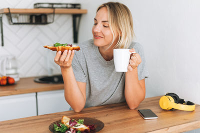 Young woman having breakfast with healthy sandwiches croissants and coffee in mug in bright kitchen 