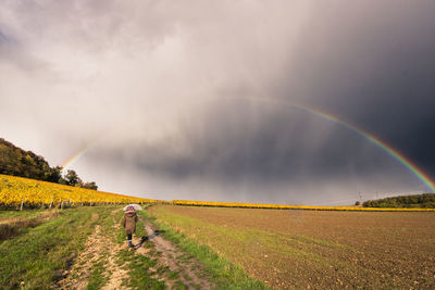 Rear view of woman holding umbrella walking on field against cloudy sky