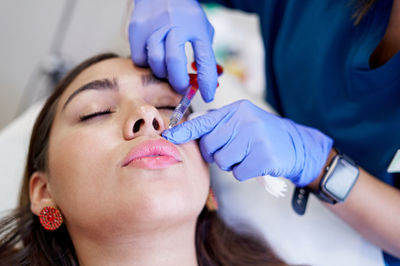 From above crop unrecognizable esthetician in latex gloves injecting collagen into lips of relaxed female client during appointment in beauty clinic