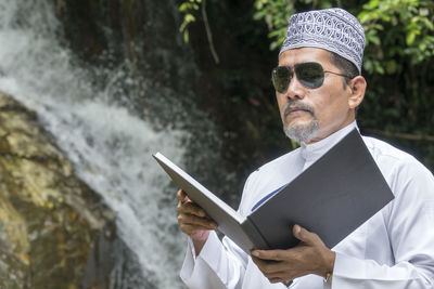 Man reading book while standing against waterfall