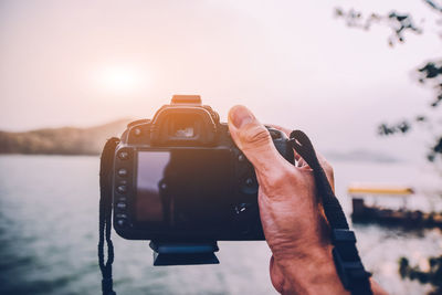 Cropped hand holding camera by lake during sunset