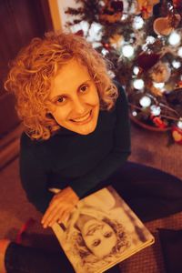 High angle portrait of smiling young woman sitting by illuminated christmas tree at home