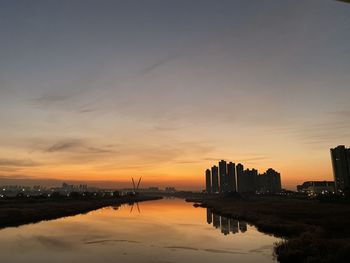 Silhouette buildings by river against sky during sunset