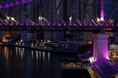 A bridge lights up in purple reflecting over the brisbane river at night.