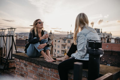 Cheerful women talking while sitting on building terrace against sky