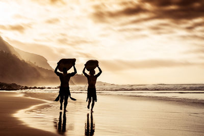 Men carrying surfboards while walking on beach against sky during sunset