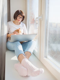 Smiling woman using laptop while sitting on window sill
