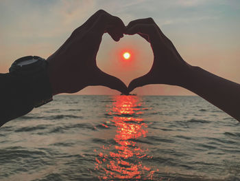 Man holding heart shape in sea against sky during sunset