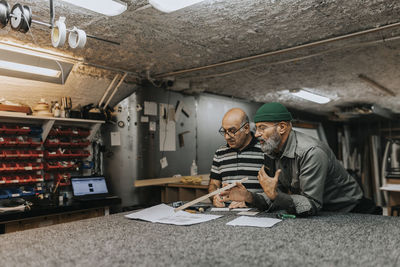 Mature craftsman discussing over blueprint with male colleague at workbench