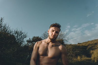Portrait of shirtless young man standing on land against sky