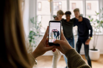 Teenage girl with smart phone filming male friends standing arm around in living room
