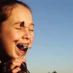 Close-up of girl with eyes closed trying to eat flower against clear blue sky