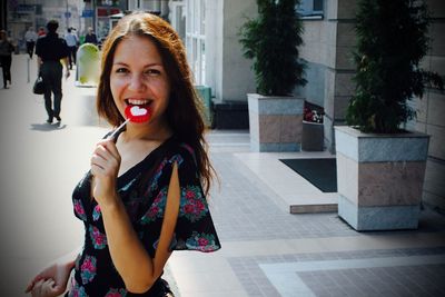 Portrait of cheerful woman eating lollipop while standing at city street