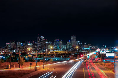 Light trails on road by illuminated buildings against sky at night