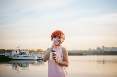 Mid adult woman talking on mobile phone while standing by river against sky during sunset