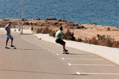 Two men playing figure skating on a rural road in the sun on a bright day play surf skate near coast