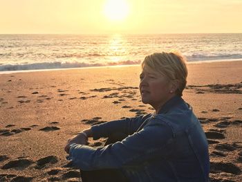 Woman sitting at beach against sky during sunset