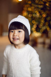 Beautiful korean child girl in in a white knitted sweater and hat stands at the christmas tree 