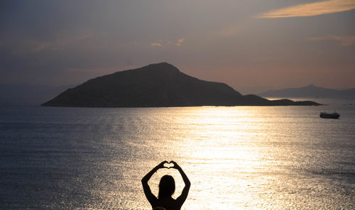 Silhouette woman forming heart shape by sea against sky during sunset