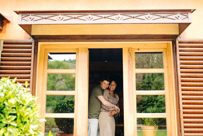 View from terrace of multiracial couple hugging while standing in room near doors of wooden house and smiling