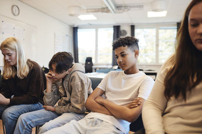 Thoughtful teenage boy sitting with arms crossed amidst friends in classroom during group therapy