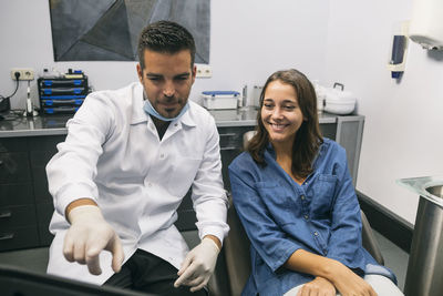 Male dentist explaining medical procedure to smiling young female patient over laptop in clinic