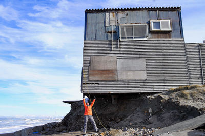 Woman standing under a derelict house holding it up at a beach in winter under blue skies 