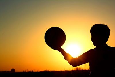 Silhouette boy holding ball against orange clear sky during sunset