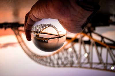 Cropped hand holding crystal ball with reflection of bridge and river during sunset