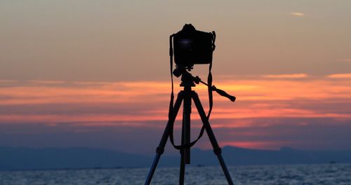 Silhouette camera on sea against orange sky