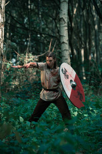 Brave female in viking costume standing with spear and shield for medieval reenactment in woods