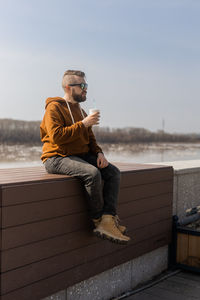 Young woman sitting on bench against sky