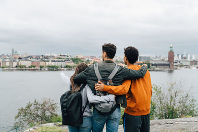 Rear view of friends with arms around standing by river in city against sky