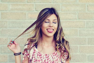 Young woman with eyes closed sticking out tongue while standing against brick wall