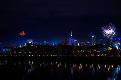 Firework display over river and illuminated buildings in city at night