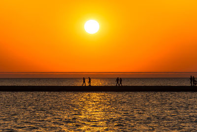 Silhouette people on sea against orange sky