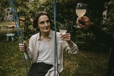 Smiling man toasting drink with friend while sitting on swing in yard