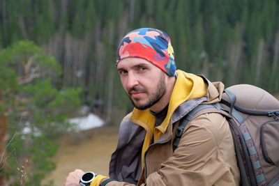 A tourist in a brown jacket and a bright camouflage bandana sits on a high bank of a mountain river. 