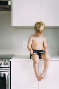 Shirtless boy using tablet computer while sitting on kitchen counter at home