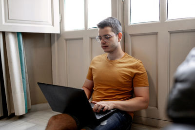 Focused male freelancer in casual outfit sitting on floor near window and typing on laptop while working remotely from home